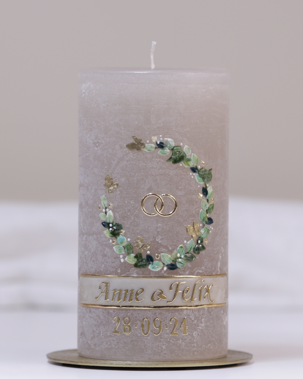 Sandgraue Hochzeitskerze mit Eheringen, grünem Blumenkranz und goldenen Schmetterlingen – detailverliebt gestaltet.