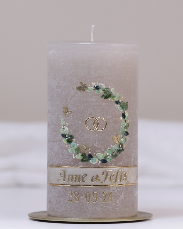 Sandgraue Hochzeitskerze mit Eheringen, grünem Blumenkranz und goldenen Schmetterlingen – detailverliebt gestaltet.